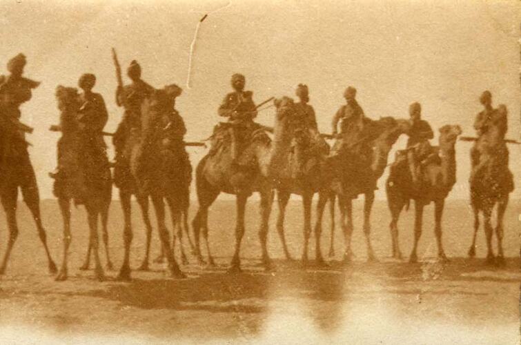 Row of men on camels wearing turbans and holding rifles. They are in the desert.