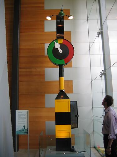 Man looking up at yellow and black striped pole with green and red round dial near top.
