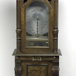 Decorative wooden upright case with glass pane door. Contains metal disc and mechanism.