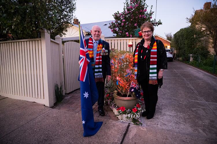 Man and woman stand in front of a picket-fenced house. He wears military medals and holds an Australian flag.