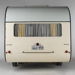 Cream caravan, curved edges, metal trim, rear view. One window, yellow white curtain. Number plate.