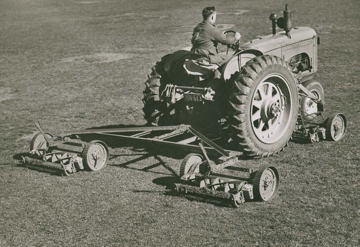 Man driving a tractor towing a cylindirical mower,  also fitted with a mid-mounted cylindrical mower.