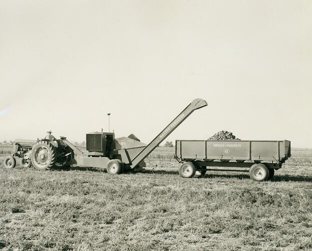 Man driving a tractor pulling a hay pelleting machine, dumping the pellets into a farm trailer.