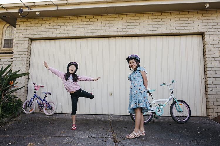 Two smiling girls pose in front of a garage with their bikes nearby. One has both her arms and one leg raised.
