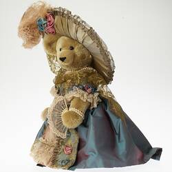 Light brown plush bear wearing lavish cream hat with feather and ornate dusty pink dress and blue grey jacket.