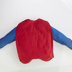 Red cape back of child's long sleeve blue top with yellow cuffs. Red cape hangs down back.