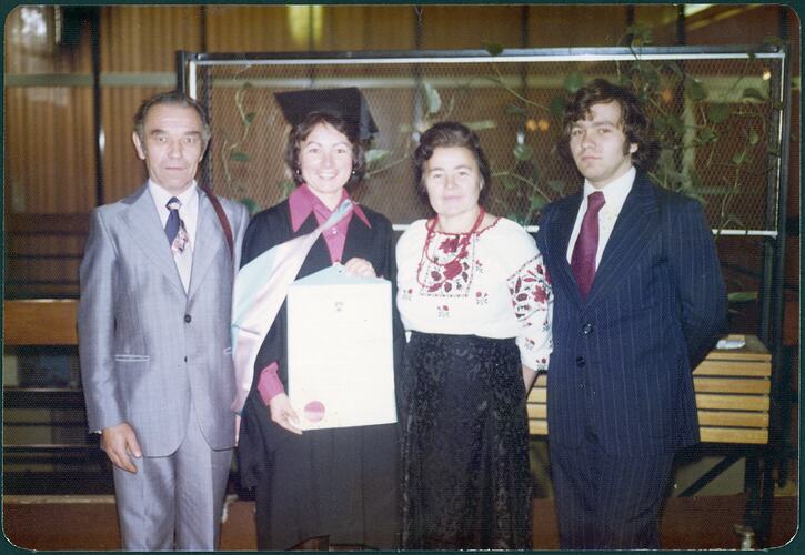 Woman posing at her graduation holding a framed certificate. Her father is at the left and her mother and brother at the right.