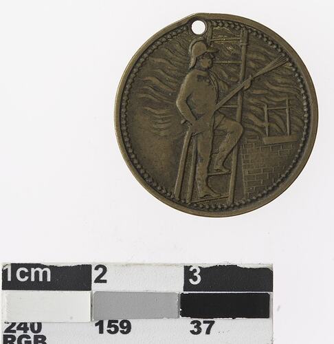 Round bronze coloured medal with man on ladder with hose against building with flames emanating.