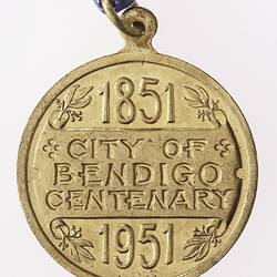 Round gold coloured medal with text in centre, blue, white and red ribbon attached.