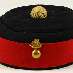 Black wool hat with red wool band and gold broach around rim and yellow decoration on top centre.