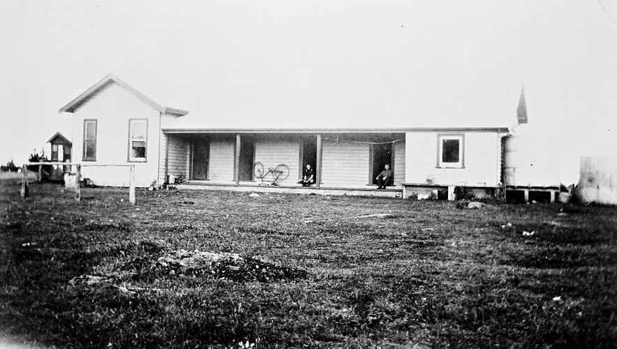 Two men seated in the doorway of a building. There is an upside-down bicycle on the verandah at the left.