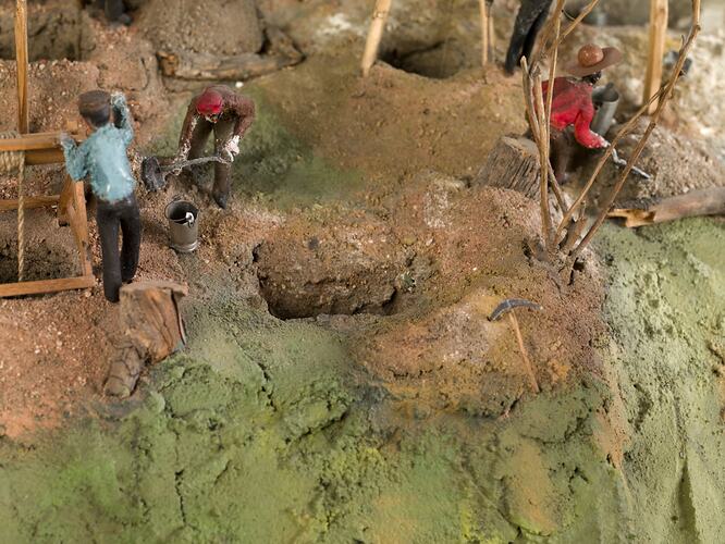 Detail of goldmine model. Three figures are working with tools on an uneven hillside with several open minesha