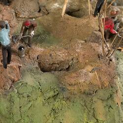 Detail of goldmine model. Three figures are working with tools on an uneven hillside with several open minesha