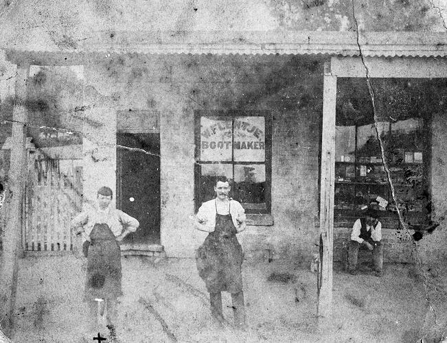 Two men wearing aprons standing in front of a bootmaker's shop. Man sits on seat at right.