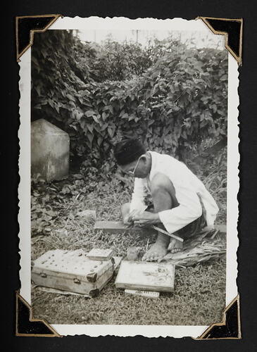 Man sitting o ground with suitcase and other objects, leafy foliage behind.