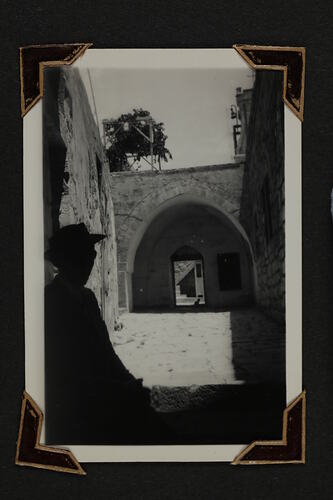 Woman in shadow in foreground, stone walkway with walls leading to arch and doorway in background.