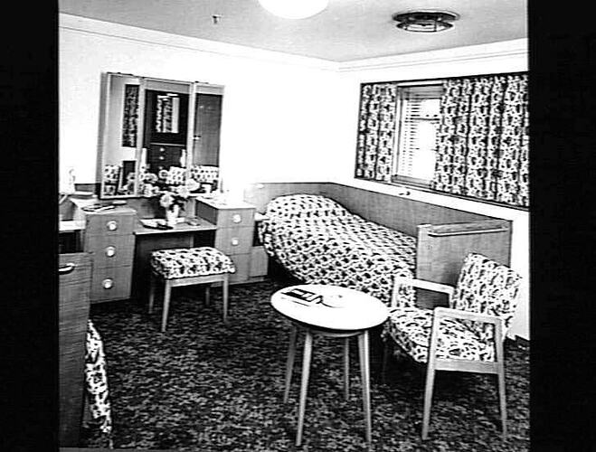 Ship interior. Single bed against wall, dressing table, chair and coffee table.