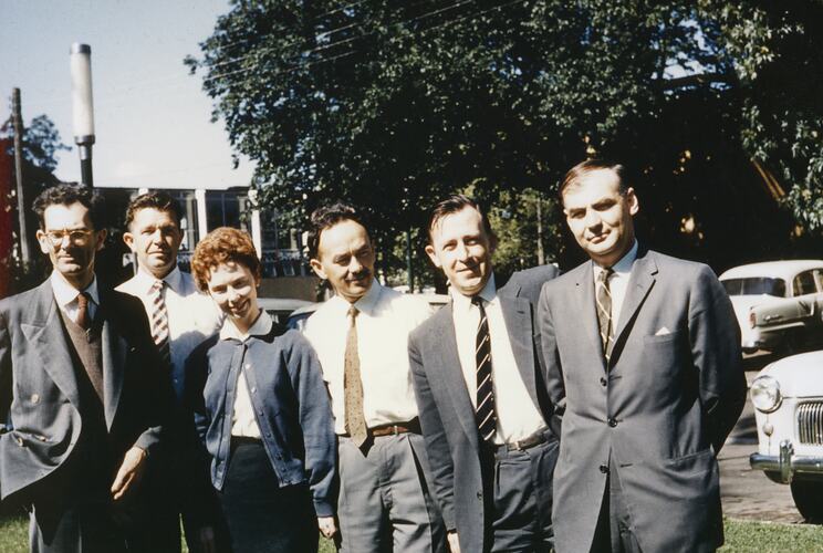 Five men and one woman pose in front of a building and trees.