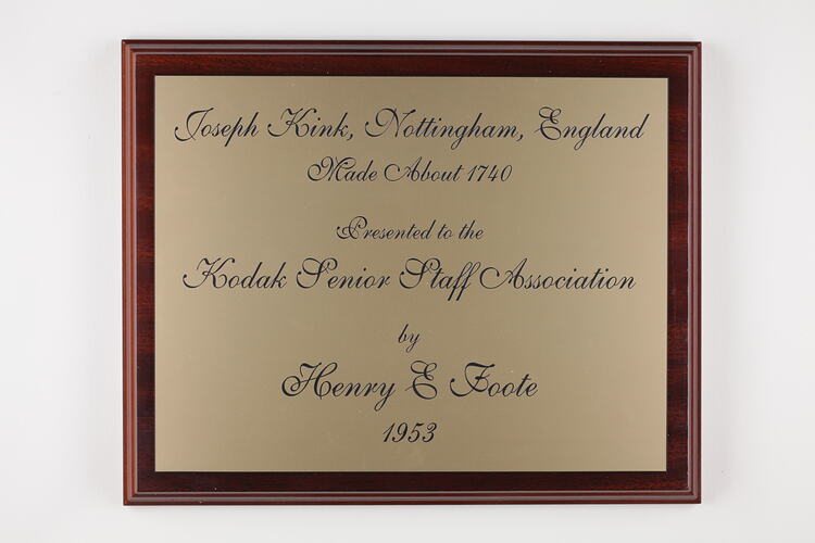 Rectangular, gold, engraved plaque mounted on wood.