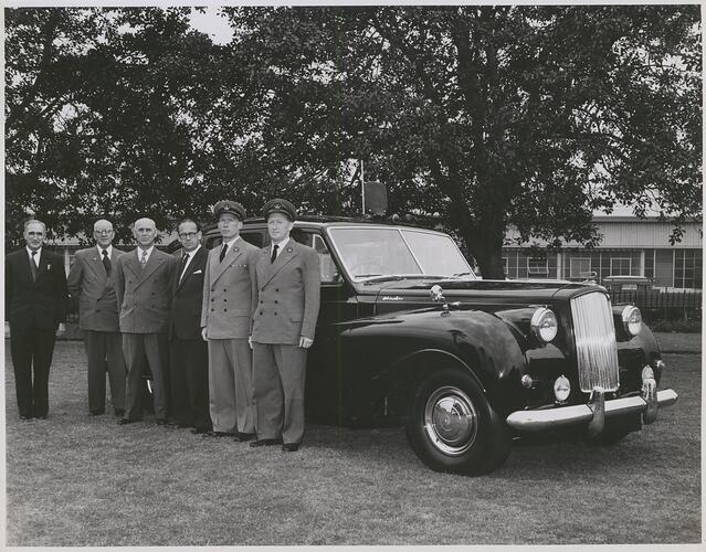 Six men in suits pose formally in front of a car.