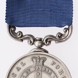 Round silver medal with ornate mount suspended from blue ribbon. Medal has crown, cross and text.