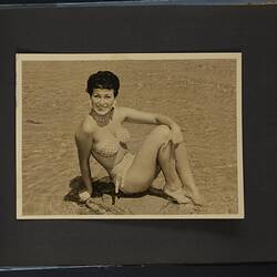 Album page, photograph, black and white, woman in polka dot bikini posing seated on beach.
