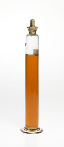 Cylindrical glass jar with amber coloured liquid. Two labels affixed, sealed at top.