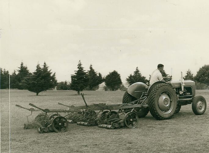 Man driving a tractor towing a cylindrical gang mower on playing field.