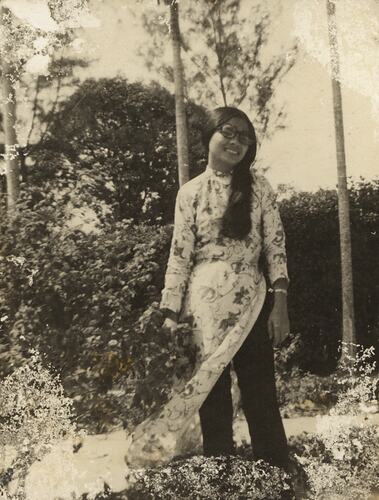 Woman with long black hair standing with shrubbery in background.