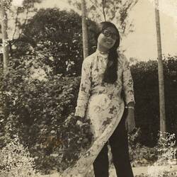 Woman with long black hair standing with shrubbery in background.