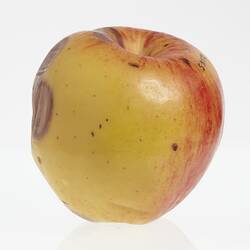 Wax model of an apple painted yellow with some red, Has brown round spots on side.