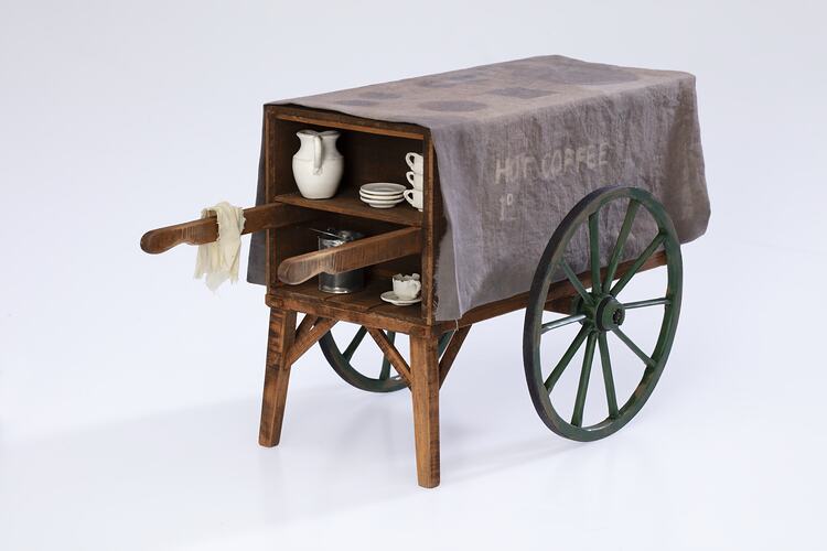 Miniature wooden coffee hand cart with two legs, two wheels. Internal shelf has coffee items, jug, cups, tin.
