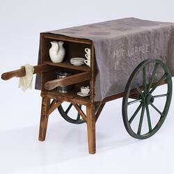 Miniature wooden coffee hand cart with two legs, two wheels. Internal shelf has coffee items, jug, cups, tin.