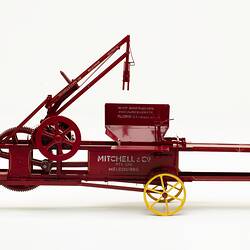 Red metal hay press model with turning gears and springs, four yellow wheels and white painted letters on side