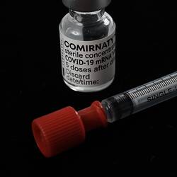 Glass vial with white printed label and metal cap behind syringe with black text and red cap.