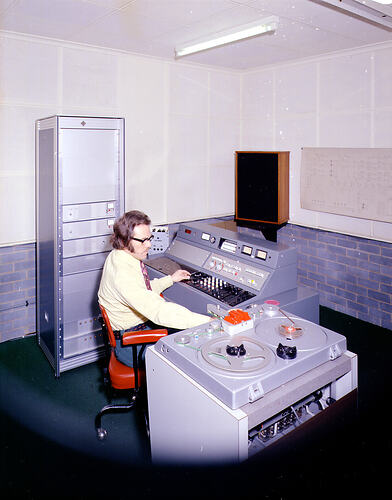 Man, seated, operating two consoles with dials, switches and tape reels.