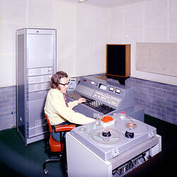 Man, seated, operating two consoles with dials, switches and tape reels.