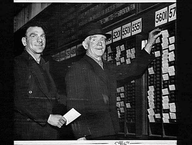 MART PHELAN SNR., (RIGHT) AND HIS SON, MART, ATTEND TO THEIR PRE-WORK DUTY AT THE TIME CARD RACKS. BOTH ARE ENGAGED IN THE SHEET IRON DEPARTMENT. MART SNR. STARTED WORK WITH H.V. MCKAY AT BALLARAT IN OCTOBER, 1899. HE IS AT PRESENT OUR OLDEST EMPLOYEE, WI
