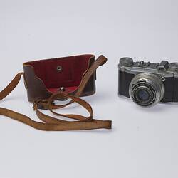 Black and silver camera. Central lens at front and two dials on top. Brown leather camera case with strap.