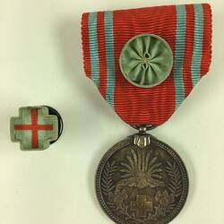 Medal suspended from red ribbon. Cross shaped pin with red cross beside it.