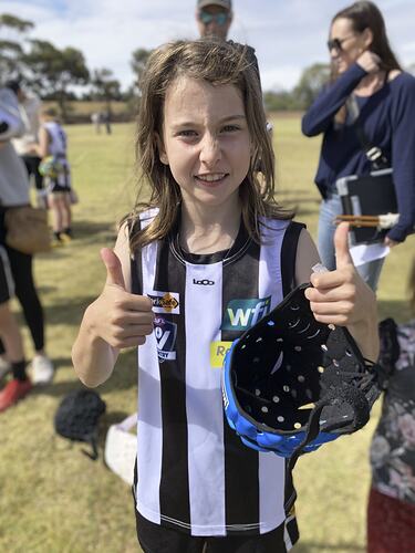 Half portrait, girl wearing black and white striped football jumper, holding her helmet and giving a double 'thumbs up'.