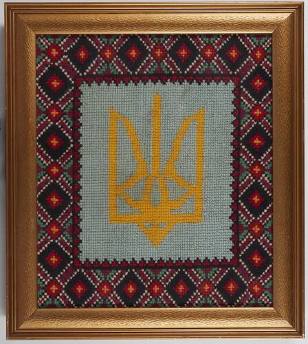 Framed tapestry featuring a yellow trident design on pale background. Dark geometric patterned embroidered border.