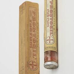 Rectangular, narrow, brown cardboard box with printing next to glass tube with metal lid. Contains red tablets. Paper labels with text affixed to tube.