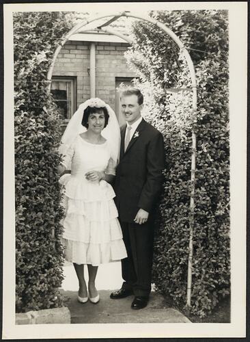 Bride and groom stand outside in garden setting under an archway.