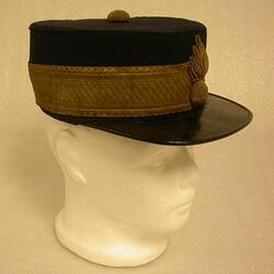 Black cap with gold hat band and leather visor on white mannequin head, side view.