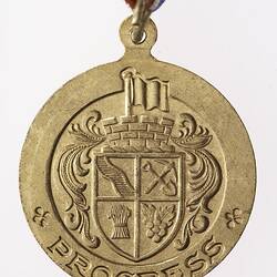 Round gold coloured medal with coat of arms, text below and red, white and blue ribbon..
