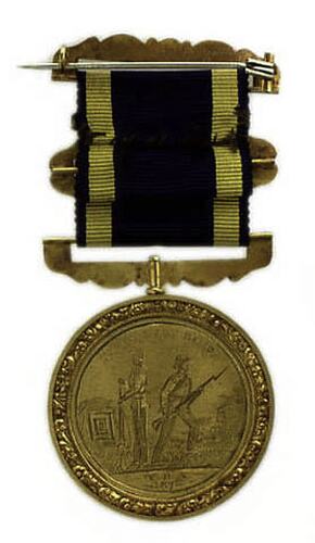Round, gold coloured medal with two men holding long rifles, blue and yellow ribbon attached.