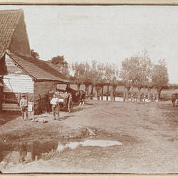 Backyard with house, two men, horse and cart on left, group of horses on right, tree lined pond in background.