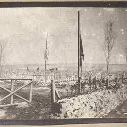Cemetery with rows of crosses and fence and gate in foreground, snow on ground.