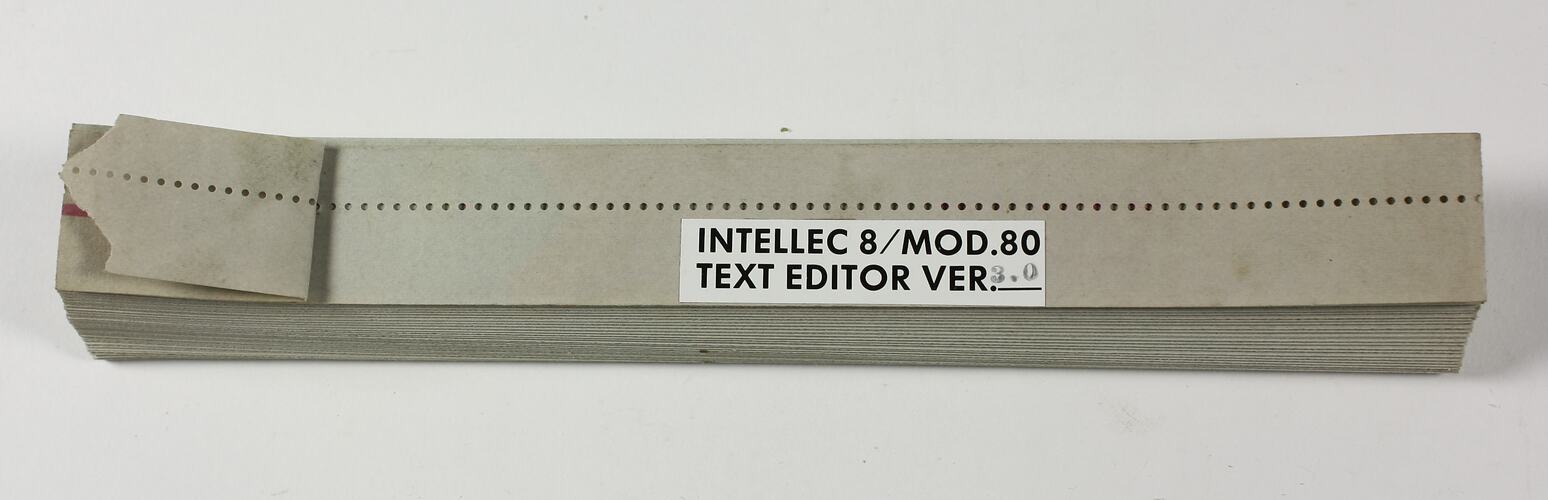 Length of neatly folded grey paper tape with punched holes. White label on top with black text.